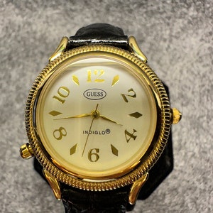 May include: A gold-tone Guess watch with a black leather strap. The watch face is white with black Roman numeral hour markers and a black Indiglo logo. The watch has a gold-tone bezel with a textured edge.
