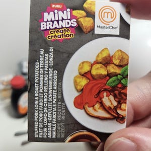 May include: A miniature food box featuring a MasterChef logo and the Mini Brands logo. The box displays an image of stuffed pork loin and roast potatoes. The text on the box includes the words "create création" and recipe instructions.