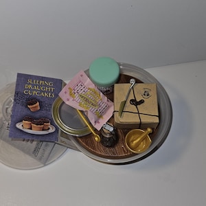 May include: A clear tray with a brown wooden base holds a variety of items, including a small book titled "Sleeping Draught Cupcakes", a pink and white box with the words "Fizzing Whizbees", a small green jar, a small brown box with a gold seal, a silver spoon, a gold mortar and pestle, and a small bottle with a cork.