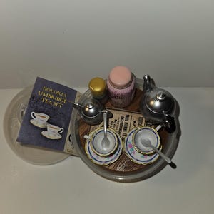 May include: A miniature tea set in a clear plastic container. The set includes a silver teapot, two teacups and saucers, a sugar bowl, a creamer, and a spoon. The teacups have a floral design. The container has a label that reads "Dolores Umbridge Tea Set".