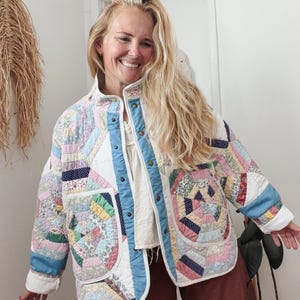 May include: A quilted jacket constructed from various patterned fabric scraps in pastel pink, blue, yellow, and white. The jacket features a high collar, a front button closure with metallic snaps, long sleeves, and two large patch pockets on the front.