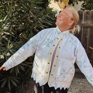 May include: A white quilted jacket featuring a repeating pinwheel pattern in soft pastel shades of green, blue, and pink. The jacket has a high collar, a front button closure with a solid cream-coloured placket, two large patch pockets, and a scalloped hem.