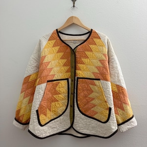 May include: A quilted jacket featuring a geometric star pattern in shades of yellow and orange. The garment has a cream-colored base, a round neckline, long sleeves, and two front patch pockets. It is finished with dark brown trim and buttons.