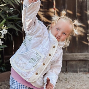 May include: A quilted jacket featuring a white base with a subtle multicolored grid pattern, a high collar, long sleeves, and a front button closure with metallic hardware. The jacket includes two patch pockets on the front and a cream-colored trim.