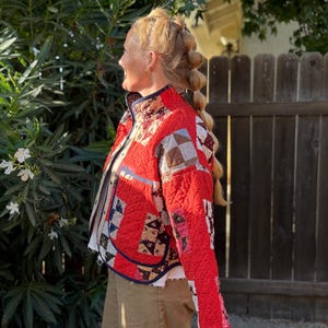 May include: A quilted jacket featuring a patchwork design with a dominant bright red color and various patterned squares in brown, white, and blue. The jacket has a high collar, long sleeves, and dark blue trim along the edges and front opening.