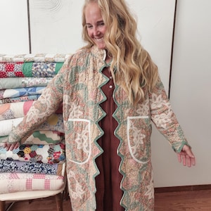 May include: A long, quilted coat featuring a floral pattern in muted shades of green, pink, and cream. The garment has a distinctive scalloped front edge, long sleeves, and two large patch pockets with white trim, worn over a solid brown dress.