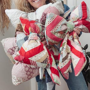 Puede incluir: Una colección de almohadas hechas a mano con forma de lazo, hechas con patchwork, en varios colores y patrones. Las almohadas presentan telas rojas, blancas, rosas y estampadas, mostrando una estética vintage. Los lazos están atados en el centro, creando un artículo decorativo único.