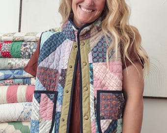 Puede incluir: Un chaleco acolchado con un diseño de patchwork de varios patrones de tela en tonos rosa, azul y verde. El chaleco tiene un cuello alto, un borde oscuro a lo largo de los contornos, dos bolsillos delanteros y una fila de botones a presión color latón.