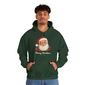 May include: A dark green hooded sweatshirt featuring a graphic of a winking Santa Claus wearing a red hat with a white pom-pom. Below the illustration, the text Merry Christmas is printed in a white, cursive, vintage-style font.