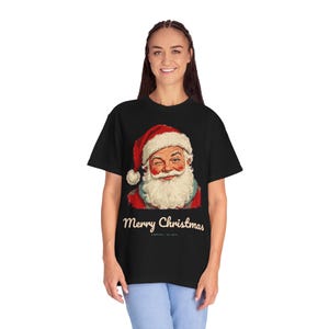 May include: A black short-sleeve t-shirt featuring a graphic of a winking Santa Claus wearing a red hat with white trim. Below the illustration, the text reads Merry Christmas in a cream-colored script font, followed by smaller text.