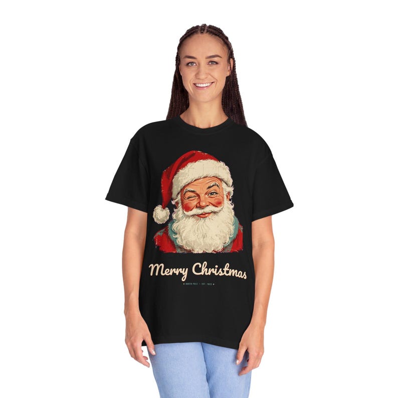 May include: A black short-sleeve t-shirt featuring a graphic of a winking Santa Claus wearing a red hat with white trim. Below the illustration, the text reads Merry Christmas in a cream-colored script font, followed by smaller text.