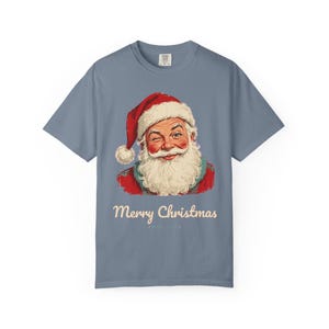 May include: A slate blue short-sleeve t-shirt featuring a graphic of a winking Santa Claus wearing a red hat with a white pom-pom. Below the illustration, the text Merry Christmas is printed in a cream-colored script font.