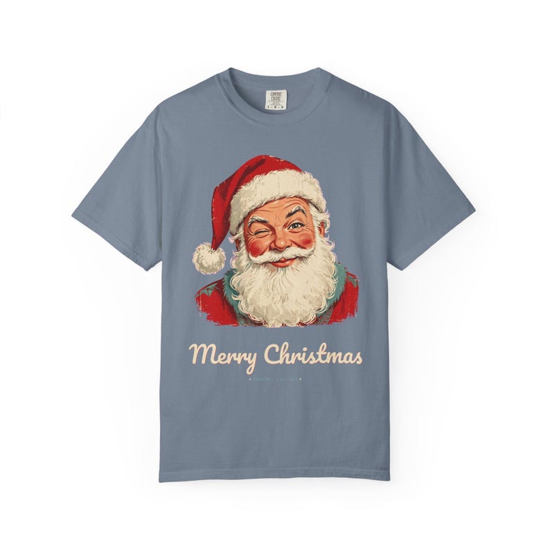 May include: A slate blue short-sleeve t-shirt featuring a graphic of a winking Santa Claus wearing a red hat with a white pom-pom. Below the illustration, the text Merry Christmas is printed in a cream-colored script font.