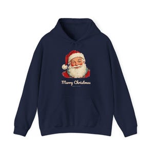 May include: A navy blue hooded sweatshirt featuring a graphic of a winking Santa Claus wearing a red hat with white trim. Below the illustration, the text Merry Christmas is printed in a cream-colored script font on the front of the garment.