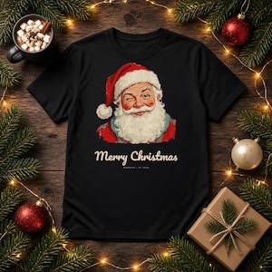 May include: A black short-sleeve t-shirt featuring a graphic of a winking Santa Claus with a white beard and red hat. Below the illustration, the text reads Merry Christmas in a cream-colored font, followed by North Pole Est. 1822.