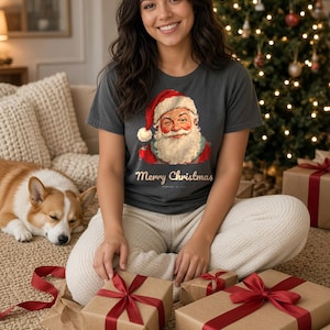 May include: A charcoal gray short-sleeve t-shirt featuring a graphic of a winking Santa Claus wearing a red hat and the text Merry Christmas in a cursive font. The shirt is paired with cream-colored knit lounge pants.