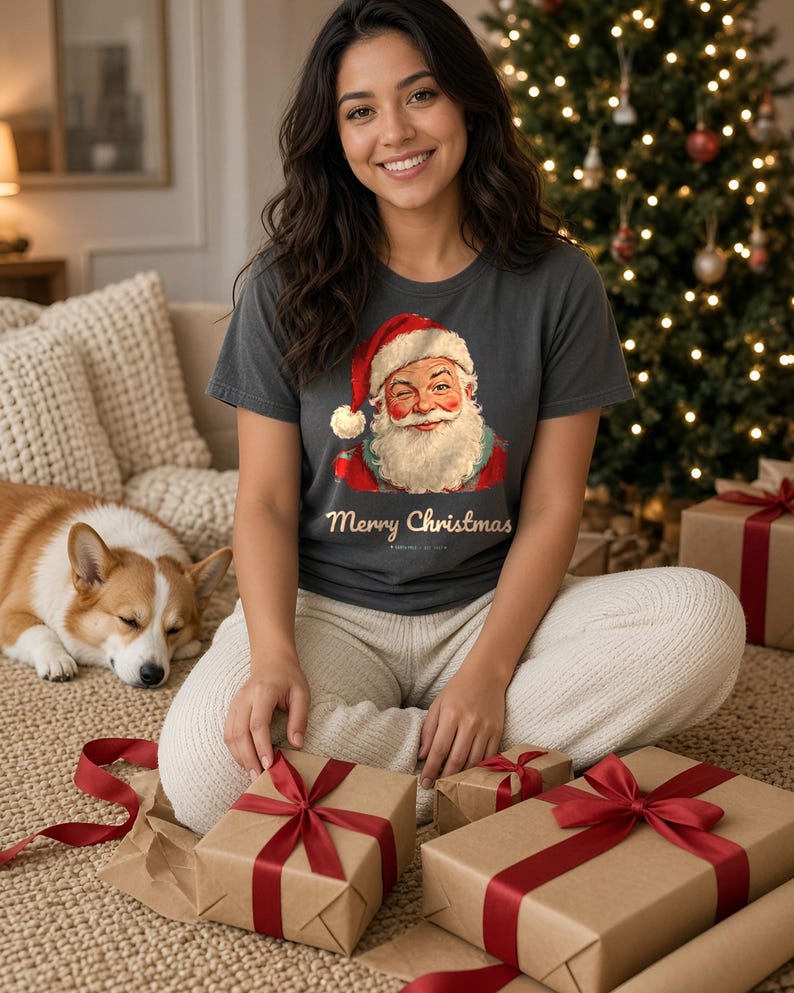May include: A charcoal gray short-sleeve t-shirt featuring a graphic of a winking Santa Claus wearing a red hat and the text Merry Christmas in a cursive font. The shirt is paired with cream-colored knit lounge pants.