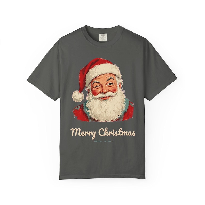 May include: A charcoal gray short-sleeve t-shirt featuring a graphic of a winking Santa Claus wearing a red hat and white beard. Below the illustration, the text reads Merry Christmas in a cream-colored script font on the front center.