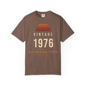 K&ouml;nnte beinhalten: Ein braunes Kurzarm-T-Shirt mit einer Grafik, die ein Sonnenuntergangsmotiv &uuml;ber dem Text VINTAGE 1976 zeigt. Unter der Jahreszahl steht der Text ALL ORIGINAL PARTS (MOSTLY) in cremefarbenen und orangefarbenen Buchstaben.