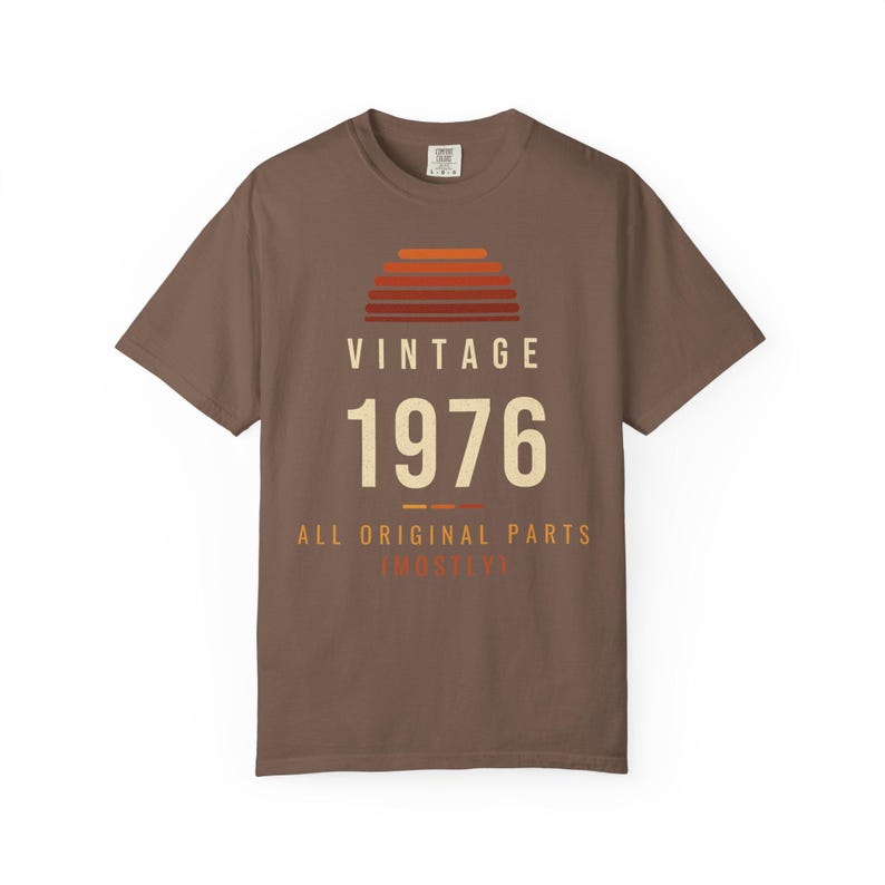K&ouml;nnte beinhalten: Ein braunes Kurzarm-T-Shirt mit einer Grafik, die ein Sonnenuntergangsmotiv &uuml;ber dem Text VINTAGE 1976 zeigt. Unter der Jahreszahl steht der Text ALL ORIGINAL PARTS (MOSTLY) in cremefarbenen und orangefarbenen Buchstaben.