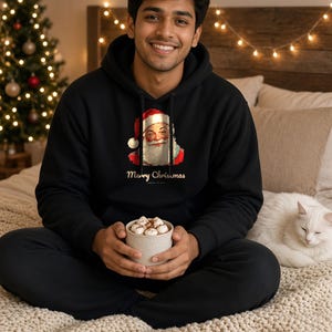 May include: A black hooded sweatshirt featuring a graphic of Santa Claus wearing a red hat and the text Merry Christmas printed in white. The hoodie has long sleeves, a front kangaroo pocket, and a drawstring hood made of cotton fabric.