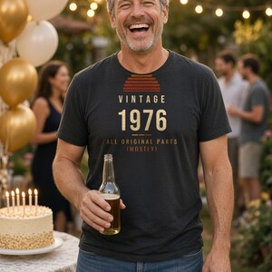 K&ouml;nnte beinhalten: Ein anthrazitfarbenes Kurzarm-T-Shirt mit einer Grafik, die ein Sonnenuntergangssymbol und den Text VINTAGE 1976 ALL ORIGINAL PARTS (MOSTLY) zeigt. Das T-Shirt hat einen Rundhalsausschnitt und besteht aus weichem, meliertem Stoff.
