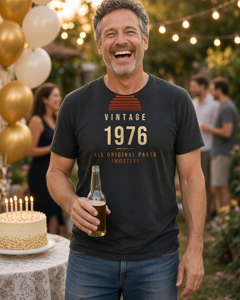 K&ouml;nnte beinhalten: Ein anthrazitfarbenes Kurzarm-T-Shirt mit einer Grafik, die ein Sonnenuntergangssymbol und den Text VINTAGE 1976 ALL ORIGINAL PARTS (MOSTLY) zeigt. Das T-Shirt hat einen Rundhalsausschnitt und besteht aus weichem, meliertem Stoff.