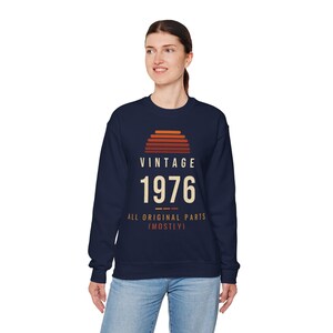以下が含まれることがあります： ネイビーブルーのクルーネックトレーナー。胸元には夕日のアイコンと「VINTAGE 1976」、そして「ALL ORIGINAL PARTS (MOSTLY)」という文字がクリーム色とオレンジ色のレタリングでプリントされています。