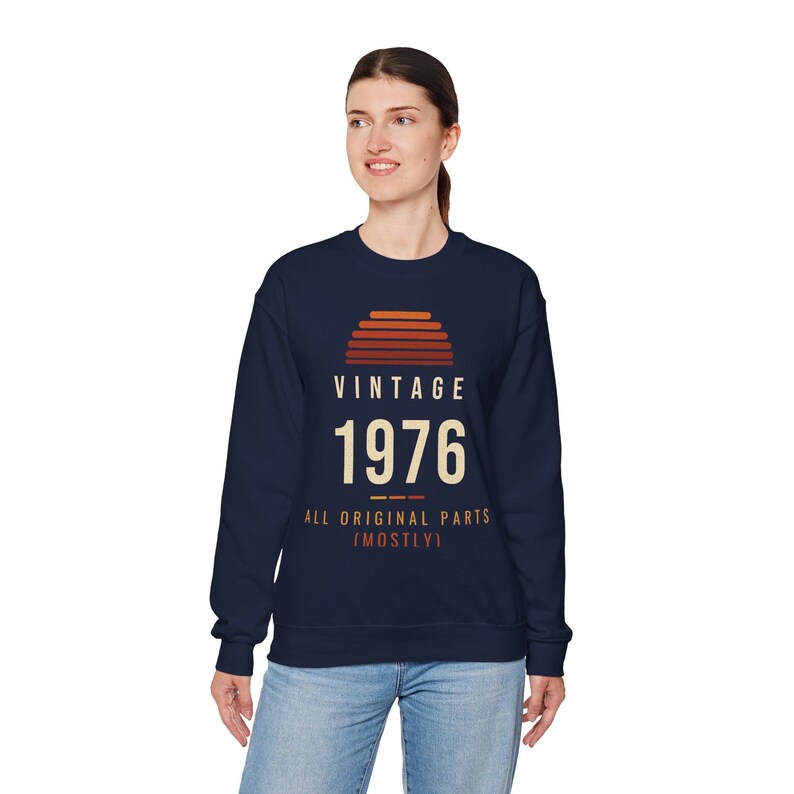 以下が含まれることがあります： ネイビーブルーのクルーネックトレーナー。胸元には夕日のアイコンと「VINTAGE 1976」、そして「ALL ORIGINAL PARTS (MOSTLY)」という文字がクリーム色とオレンジ色のレタリングでプリントされています。