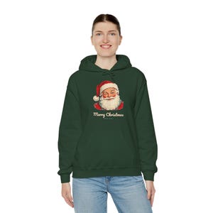 May include: A dark green hooded sweatshirt featuring a graphic of a winking Santa Claus wearing a red hat and the text Merry Christmas printed in a cream-colored font centered on the chest area of the garment.