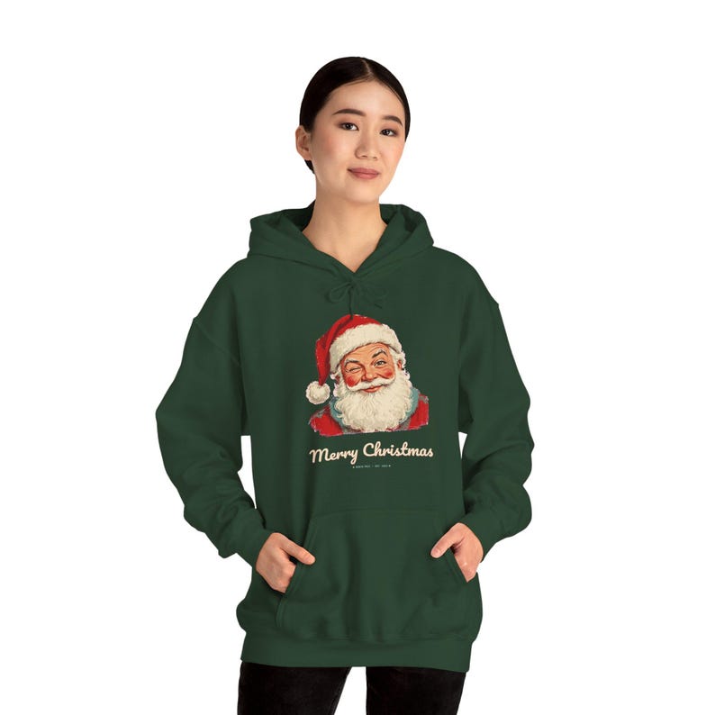 May include: A dark green hooded sweatshirt featuring a graphic of a winking Santa Claus wearing a red hat and white beard. Below the illustration, the text Merry Christmas is printed in a cream-colored script font on the front center.