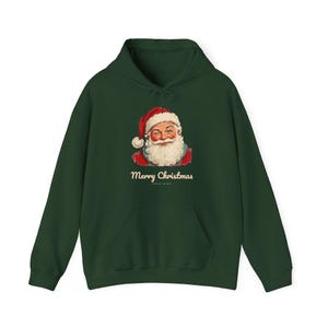 May include: A dark green hooded sweatshirt featuring a graphic of a winking Santa Claus wearing a red hat and white beard. Below the illustration, the text Merry Christmas is printed in a cream-colored script font on the front.