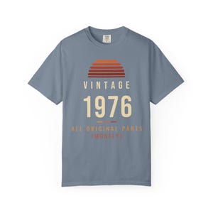 K&ouml;nnte beinhalten: Ein schieferblaues Kurzarm-T-Shirt mit einer Grafik eines Sonnenuntergangs in Orange- und Rott&ouml;nen. Der Text VINTAGE 1976 ALL ORIGINAL PARTS (MOSTLY) ist in cremefarbenen und orangefarbenen Buchstaben auf der Vorderseite abgebildet.