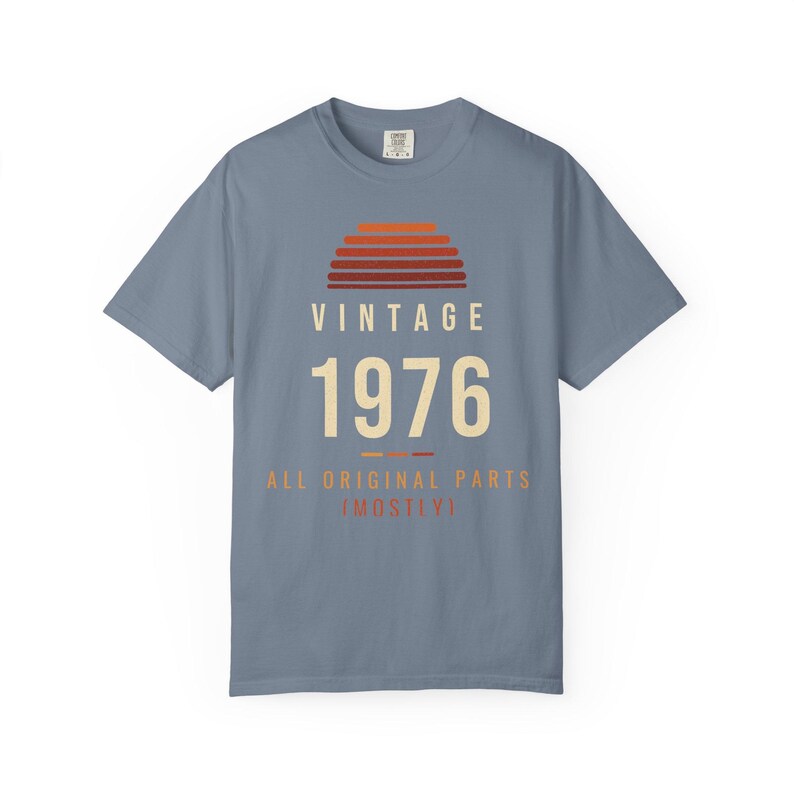 K&ouml;nnte beinhalten: Ein schieferblaues Kurzarm-T-Shirt mit einer Grafik eines Sonnenuntergangs in Orange- und Rott&ouml;nen. Der Text VINTAGE 1976 ALL ORIGINAL PARTS (MOSTLY) ist in cremefarbenen und orangefarbenen Buchstaben auf der Vorderseite abgebildet.