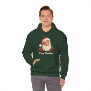 May include: A dark green hooded sweatshirt featuring a graphic of Santa Claus wearing a red hat and white beard. Below the illustration, the text reads Merry Christmas in a cream-colored font. The hoodie includes a front kangaroo pocket.