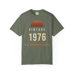K&ouml;nnte beinhalten: Ein olivgr&uuml;nes Kurzarm-T-Shirt mit einer Grafik eines Sonnenuntergangs aus horizontalen Streifen in Orange- und Rott&ouml;nen. Unter dem Sonnenuntergang steht der Text VINTAGE 1976 ALL ORIGINAL PARTS in cremefarbener und oranger Schrift.