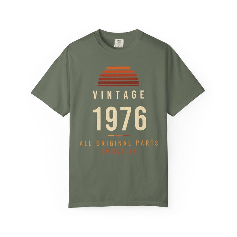 K&ouml;nnte beinhalten: Ein olivgr&uuml;nes Kurzarm-T-Shirt mit einer Grafik eines Sonnenuntergangs aus horizontalen Streifen in Orange- und Rott&ouml;nen. Unter dem Sonnenuntergang steht der Text VINTAGE 1976 ALL ORIGINAL PARTS in cremefarbener und oranger Schrift.