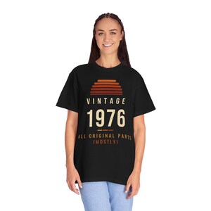 K&ouml;nnte beinhalten: Ein schwarzes Kurzarm-T-Shirt mit einer Grafik, die einen Sonnenuntergang in Orange- und Braunt&ouml;nen zeigt. Der Text VINTAGE 1976 ALL ORIGINAL PARTS (MOSTLY) ist in cremefarbenen und orangen Buchstaben auf der Vorderseite abgebildet.