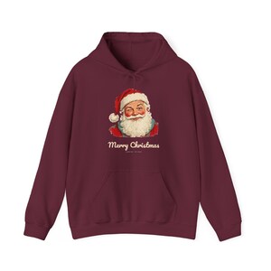May include: A deep burgundy hooded sweatshirt featuring a graphic of a winking Santa Claus wearing a red hat and white beard. Below the illustration, the text Merry Christmas is printed in a cream-colored script font on the front.