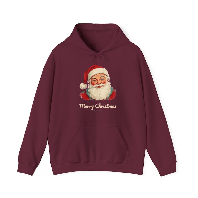 May include: A deep burgundy hooded sweatshirt featuring a graphic of a winking Santa Claus wearing a red hat and white beard. Below the illustration, the text Merry Christmas is printed in a cream-colored script font on the front.