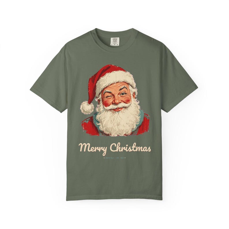 May include: An olive green short-sleeve t-shirt featuring a graphic of a winking Santa Claus wearing a red hat with white trim. Below the illustration, the text reads Merry Christmas in a cream-colored script font on the front of the shirt.
