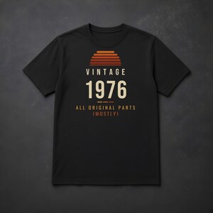 K&ouml;nnte beinhalten: Ein schwarzes Kurzarm-T-Shirt mit einem Grafikdruck in Form eines Sonnenuntergangs in Orange- und Rott&ouml;nen. Der Text VINTAGE 1976 ALL ORIGINAL PARTS (MOSTLY) ist in wei&szlig;er und oranger Schrift mittig auf der Brust platziert.