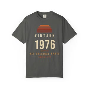 K&ouml;nnte beinhalten: Ein anthrazitfarbenes Kurzarm-T-Shirt mit einer Grafik eines Sonnenuntergangs in Orange- und Rott&ouml;nen &uuml;ber dem Text VINTAGE 1976 und dem Schriftzug ALL ORIGINAL PARTS (MOSTLY) in Creme und Orange auf der Vorderseite.