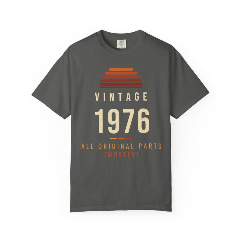 K&ouml;nnte beinhalten: Ein anthrazitfarbenes Kurzarm-T-Shirt mit einer Grafik eines Sonnenuntergangs in Orange- und Rott&ouml;nen &uuml;ber dem Text VINTAGE 1976 und dem Schriftzug ALL ORIGINAL PARTS (MOSTLY) in Creme und Orange auf der Vorderseite.