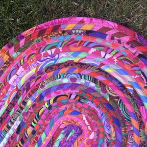 May include: A colorful round rug made of braided fabric strips in shades of pink, purple, orange, and green. The rug is made in a concentric circle pattern with a variety of floral and geometric designs.