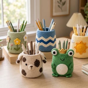 Könnte beinhalten: Eine Sammlung von fünf zylindrischen Stiftehaltern aus Häkelarbeit in verschiedenen Designs, darunter ein grüner mit Blumen, ein blauer mit Wellen, ein gelber mit Sonne, ein beiger mit braunen Punkten und ein grüner in Froschform.
