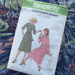 May include: Simplicity Contemporary Fashion sewing pattern, size 12, featuring illustrations of two women in dresses. The pattern cover is white and green, with text including "Contemporary Fashion" and measurements of 87cm bust. The background is a dark blue fabric with a floral pattern.