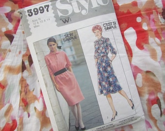May include: A vintage sewing pattern labeled "Style 3997" with illustrations of two dresses. The pattern includes sizes 10, 12, and 14. The background features a colorful floral fabric.