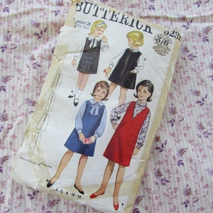 Może przedstawiać: Vintage wzór szycia Butterick z ilustracjami sukienek dla dziewczynek. Wzór zawiera projekty sukienki w kratę, czarnej sukienki, niebieskiej sukienki i czerwonego kombinezonu. Wzór jest dla obwodu klatki piersiowej 71 cm.