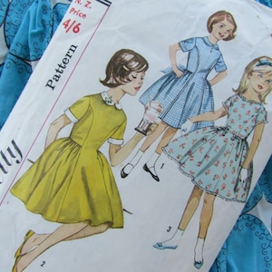May include: Vintage Simplicity sewing pattern for girls' dresses. The pattern includes three different styles: a yellow dress with white trim, a blue gingham dress with white trim, and a floral print dress with white trim. The pattern number is 4/6.